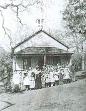 A very old photograph of a schoolhouse with students standing out front